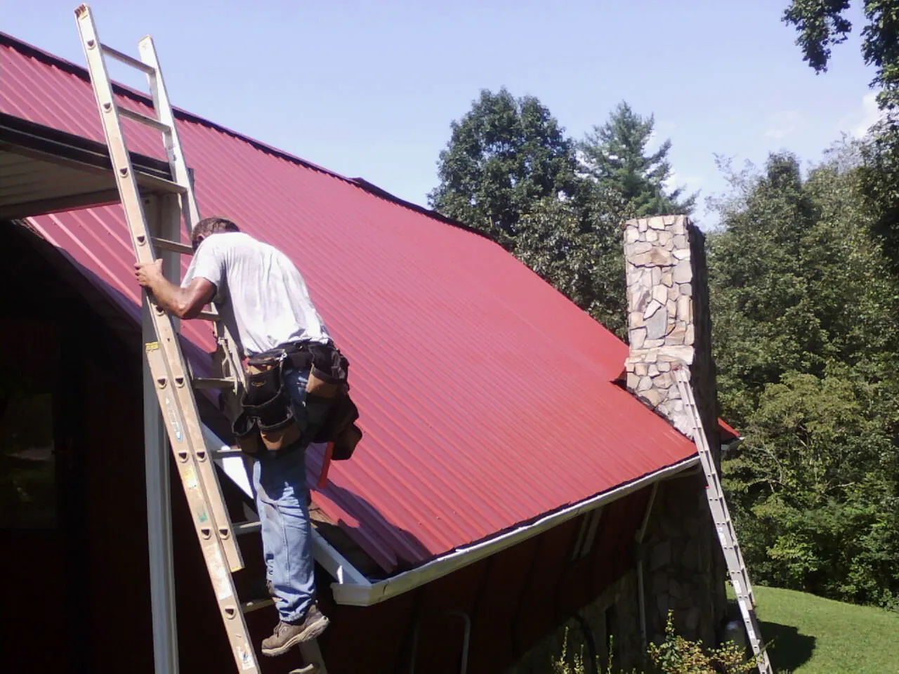 Roofer on a ladder beside a red metal roof and stone chimney — Soffit & Fascia Repair by Roofing Brothers in Penn Laird